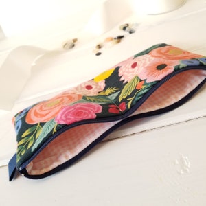 May include: A floral print pencil case with a navy blue zipper. The case features a dark background with pink, peach, and yellow flowers. The interior lining is a light pink and white houndstooth pattern.