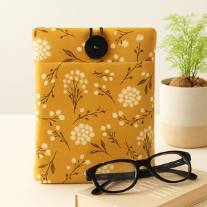May include: Mustard yellow book sleeve with a floral pattern, a black button closure, and a front pocket. Black-framed glasses rest on a book in front of the sleeve. A small potted plant is to the right.