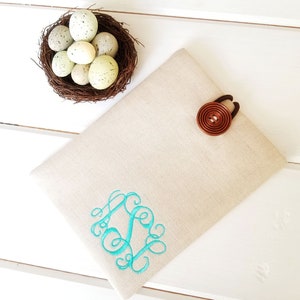 May include: A beige linen tablet sleeve with a turquoise monogram and a brown button closure. The sleeve is lying on a white wooden surface.
