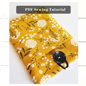 Könnte beinhalten: Eine gelbe Stofftasche mit einem weißen Blumenmuster und einem schwarzen Knopfverschluss. Die Tasche ist in der Mitte gefaltet und der Knopf ist sichtbar. Der Text "PDF Sewing Tutorial" befindet sich oben im Bild.