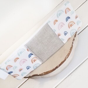 May include: A white zippered pouch with a patchwork design. One side features a light gray fabric, while the other side has a white background with a repeating pattern of watercolor rainbows in shades of pink, blue, and orange.