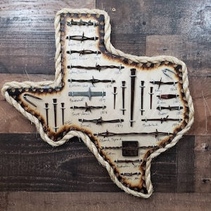 Op de afbeelding: Een verbrand houten omtrek van de staat Texas met een touwrand.  De omtrek is versierd met prikkeldraad en spijkers uit verschillende jaren.  De jaren variëren van 1877 tot 1885.  De namen van de prikkeldraad- en spijkerfabrikanten staan ​​naast elk stuk.