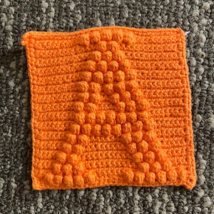 May include: A vibrant orange crocheted square, featuring a raised "A" in the centre. The handmade item displays a detailed stitch pattern, highlighting the texture and design of the craft. The background is a neutral, textured surface.
