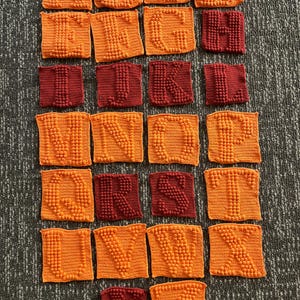 May include: A collection of crocheted alphabet squares in orange and burgundy. Each square features a raised letter, with the letters A through Z represented. The squares are arranged on a textured gray surface.