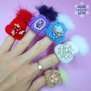 May include: Five finger rings with miniature knitted hats with different colored pom poms and embellishments. The hats are decorated with rhinestones, pearls, and other decorative elements. The rings are worn on a hand.