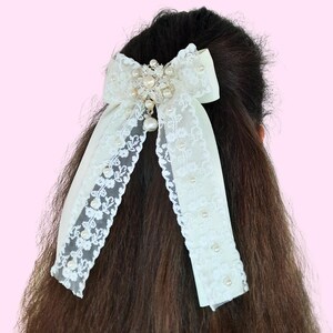 May include: A white satin bow with pearl accents and a large pearl and rhinestone embellishment. The bow is tied in a simple knot and is worn in the hair.
