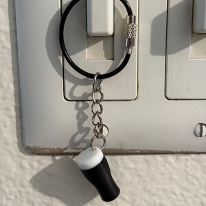 Pu&ograve; includere: Un portachiavi a forma di bicchiere da pinta in miniatura bianco e nero, con una catena d'argento e un anello nero. Il portachiavi &egrave; attaccato a un interruttore della luce. Il bicchiere da pinta ha una parte superiore bianca e un corpo nero.