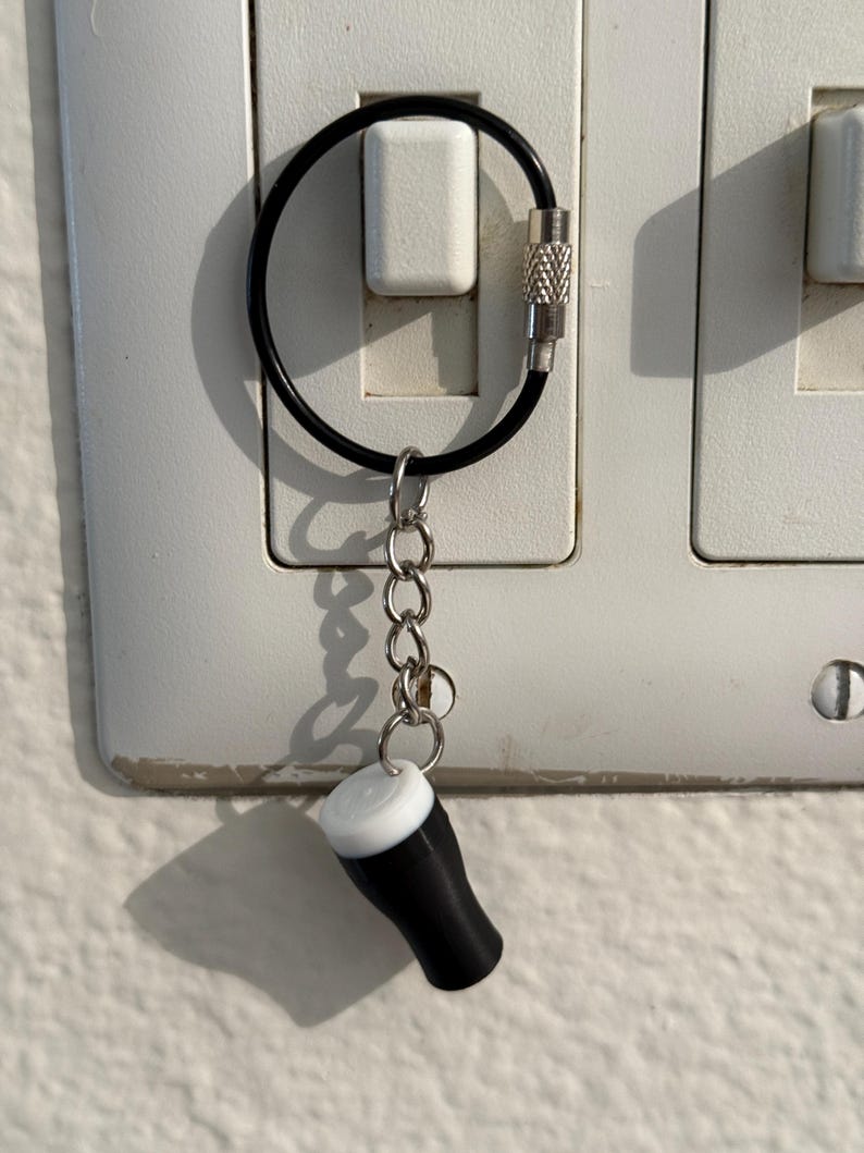 Pu&ograve; includere: Un portachiavi a forma di bicchiere da pinta in miniatura bianco e nero, con una catena d'argento e un anello nero. Il portachiavi &egrave; attaccato a un interruttore della luce. Il bicchiere da pinta ha una parte superiore bianca e un corpo nero.