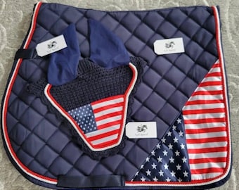 May include: A navy blue saddle pad with a white and red American flag design. The saddle pad has a white and red trim and a white label with the text "Lil' Sport" on it. The saddle pad is paired with a navy blue and white crochet ear bonnet with a red, white, and blue American flag design.