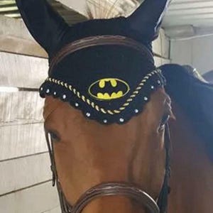 以下が含まれることがあります： 前面に黄色と黒のバットマンロゴがあしらわれた黒い馬用イヤーネット。縁はスカラップ状になっており、金色のツイストロープと小さな銀色のスタッズで装飾されています。馬の耳と額を覆うように設計されています。