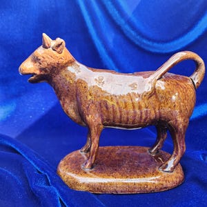 Puede incluir: Figura de vaca de cerámica marrón con cola curva y base pequeña. La vaca tiene un acabado brillante y está de pie. La figura mide aproximadamente 15 cm de largo.