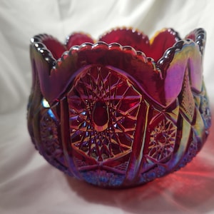 Op de afbeelding: Een robijnrode, iriserende glazen kom met een geschulpte rand. De kom heeft een gedetailleerd, geometrisch patroon met stervormige en diamantvormen. Het glas heeft een glanzende, veelkleurige afwerking.