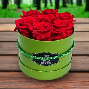 May include: A circular, lime green box overflowing with bright red roses. The box is decorated with a green rope trim and a black and white label reading "Miara Roses". The arrangement rests on a wooden surface, with a blurred forest backdrop.