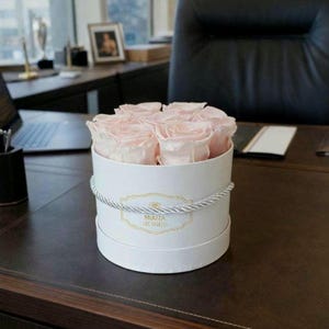 Può includere: Una scatola rotonda bianca con un dettaglio di corda argentata contiene diverse rose rosa chiaro. La scatola ha un'etichetta dorata con la scritta "MIARA ROSES". La composizione è su una superficie marrone scuro.