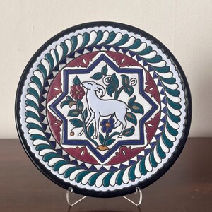 May include: A round ceramic decorative plate featuring a central white goat illustration surrounded by floral motifs within an eight-pointed star. The border displays a repeating teal and white leaf pattern, finished with a dark blue outer rim.