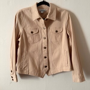 May include: A light peach-colored jacket with a vertical ribbed texture. The jacket has a collared neckline, two chest pockets, and two side pockets. It features brown buttons and long sleeves. The jacket is hanging on a black hanger.