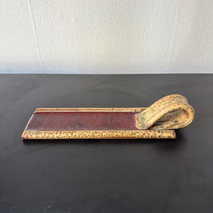 May include: A rectangular ceramic tray with a burgundy centre and a decorative, curled handle. The tray has a textured, speckled beige and brown finish with a patterned border and green accents. The tray is designed for serving or display.