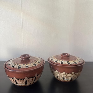 Puede incluir: Dos cuencos de cerámica marrón con tapas. Las tapas y los cuencos tienen diseños decorativos en crema y negro. Los cuencos están sobre una superficie oscura, con una pared clara al fondo.