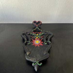 May include: Black cast iron trivet with a heart-shaped handle. The trivet features a central starburst design with red, yellow, and green accents. The decorative piece has ornate scrollwork and a textured surface.