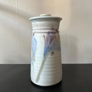 May include: A handmade ceramic jar with a lid. The jar is primarily white with blue and purple abstract designs. The jar has a cylindrical shape with a slightly wider base and a lid that fits snugly on top. The jar is approximately 6 inches tall.