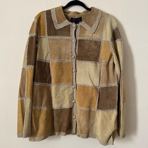 Könnte beinhalten: Eine Wildlederjacke mit Knopfleiste in Patchwork-Design. Die Jacke hat Quadrate in verschiedenen Braun-, Beige- und Beigetönen mit dekorativen Nähten. Der Kragen und die Kanten haben eine Häkelborte. Die Jacke hängt an einem schwarzen Kleiderbügel.