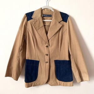 May include: Tan blazer with navy blue denim shoulder patches and pockets. The blazer has a notched collar, three buttons, and the text "tour de force" on the label.