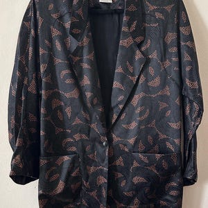 May include: A black blazer with a copper-colored swirling pattern and dotted accents. The garment features a single-button closure, notched lapels, two front pockets, and long sleeves. A label inside the collar reads PANTHER.