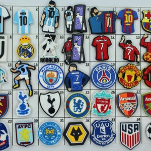 May include: A collection of 35 colorful rubber shoe charms featuring various soccer team logos, including Juventus, Real Madrid, Manchester United, Paris Saint-Germain, Arsenal, and more. The charms are arranged in a grid on a white background.