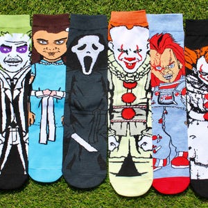 May include: Six pairs of novelty socks featuring characters from horror movies. The socks display images of Beetlejuice, Chucky, Pennywise, and the Scream mask. The socks are in various colors, including black, green, blue, and red.