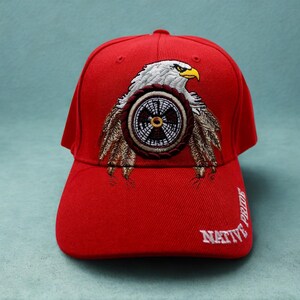 May include: A bright red baseball cap featuring an embroidered design of a white-headed eagle with a yellow beak, positioned above a circular brown and black emblem with feathers. The text NATIVE PRIDE is embroidered in white on the lower right side.