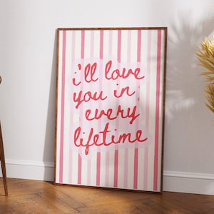May include: A framed art print with a pink and white striped background. The print features the phrase "I'll love you in every lifetime" in red script. The frame is a dark brown colour.