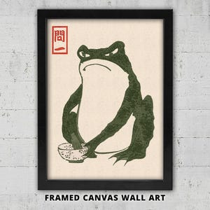 Könnte beinhalten: Gerahmtes Leinwandbild mit einer grünen Frosch-Illustration, der einen grimmigen Gesichtsausdruck hat und Stäbchen über einer Schüssel hält. Das Kunstwerk hat einen minimalistischen Stil mit japanischem Text in der oberen linken Ecke. Der Rahmen ist schwarz.