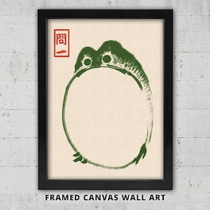 Könnte beinhalten: Gerahmtes Leinwandbild mit einer grünen froschartigen Kreatur in einer kreisförmigen Form. Das Kunstwerk hat einen minimalistischen Stil mit japanischem Text in der oberen linken Ecke. Die Leinwand ist in einem schwarzen Rahmen gerahmt.