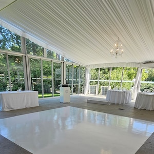 May include: An empty event space with a white dance floor, tables, and a podium. The room has large glass windows and a white fabric ceiling. A chandelier hangs from the ceiling.