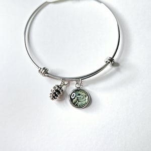May include: A silver bangle bracelet with two charms. One charm is a silver pine cone and the other is a round glass charm with a green and white design.