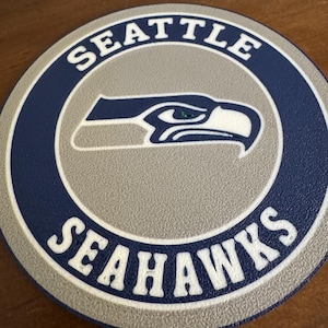 Puede incluir: Un posavasos redondo, gris y azul con las palabras "Seattle Seahawks" en letras blancas alrededor del borde. Una silueta blanca de una cabeza de halcón está en el centro.