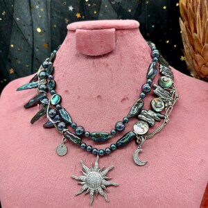 以下が含まれることがあります： 黒と虹色のバロックパール、アワビの貝殻のアクセント、太陽と月のペンダントを含むシルバーチャームがついた、重ね付けのネックレスです。