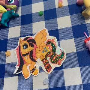 May include: A collection of My Little Pony toys and stickers on a blue and white checkered surface. The toys include a yellow pony with pink hair and wings, and a teal pony with a purple mask. A sticker of a pony with orange and black hair is also present.
