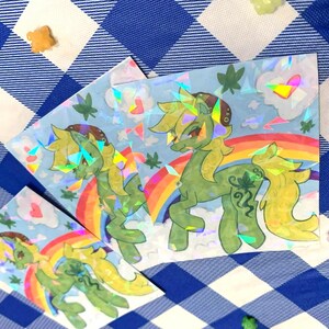 May include: Holographic stickers featuring a green pony with yellow hair, a rainbow, and a leafy design. The stickers have a light blue background with clouds and leaves. The stickers are on a blue and white checkered surface.