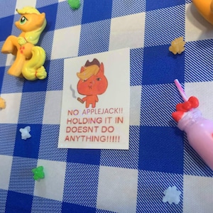 May include: A still life featuring a cartoon pony figurine, a small drawing with text, and a pink drink with a bow. The drawing says "NO APPLEJACK!! HOLDING IT IN DOESNT DO ANYTHING!!!!" on a blue and white checkered surface.