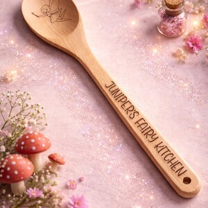 May include: Wooden spoon with a fairy illustration and the text "Juniper's Fairy Kitchen". The spoon is surrounded by pink flowers, miniature mushrooms, and a small jar with pink contents. The background is a light pink colour.