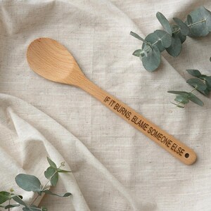 May include: A light-coloured wooden spoon with a smooth, rounded bowl and a long handle. The handle features the laser-engraved text IF IT BURNS, BLAME SOMEONE ELSE in a clean, sans-serif font, with a small circular hole at the very end.