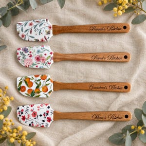 May include: Four silicone spatulas with wooden handles. Each handle is engraved with text: Nonna's Kitchen, Nana's Kitchen, Grandma's Kitchen, and Mum's Kitchen. The silicone heads feature floral and orange fruit patterns.