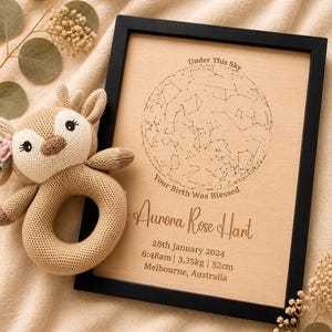 May include: A framed wooden plaque with a black border, featuring a star map and the text "Under This Sky" and "Your Birth Was Blessed". Below, it reads "Aurora Rose Hart", with birth details: 28th January 2024, 6:48am, 3.35kg, 52cm. A knitted deer rattle is next to the frame.
