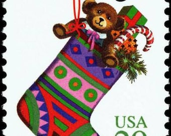 Pode incluir: Um selo postal de Natal vintage que mostra uma meia colorida com um urso de peluche dentro. O selo diz "Greetings" e "USA 29".