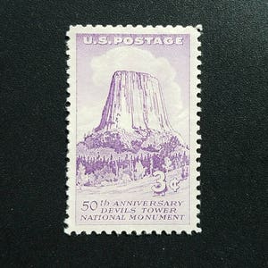 Könnte beinhalten: Eine violette und weiße US-Briefmarke zum 50. Jahrestag des Devils Tower National Monument. Die Marke zeigt eine detaillierte Illustration des Devils Tower, einer imposanten Felsformation, mit einem violetten "3¢" Wert im unteren rechten Eck.