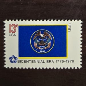 May include: A rectangular postage stamp featuring the blue flag of Utah with its central seal, an eagle, and flags. The stamp displays the text 13c USA, Utah, and BICENTENNIAL ERA 1776-1976, along with a small blue star-shaped logo on a white background.