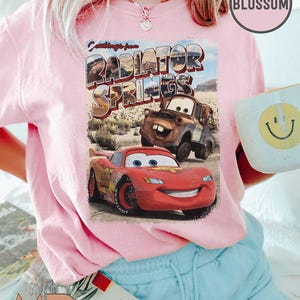 K&ouml;nnte beinhalten: Ein pinkfarbenes T-Shirt mit einem Vintage-Postkarten-Design mit dem Text "Greetings from Radiator Springs". Die Postkarte zeigt ein rotes Rennwagen und einen rostigen Abschleppwagen.
