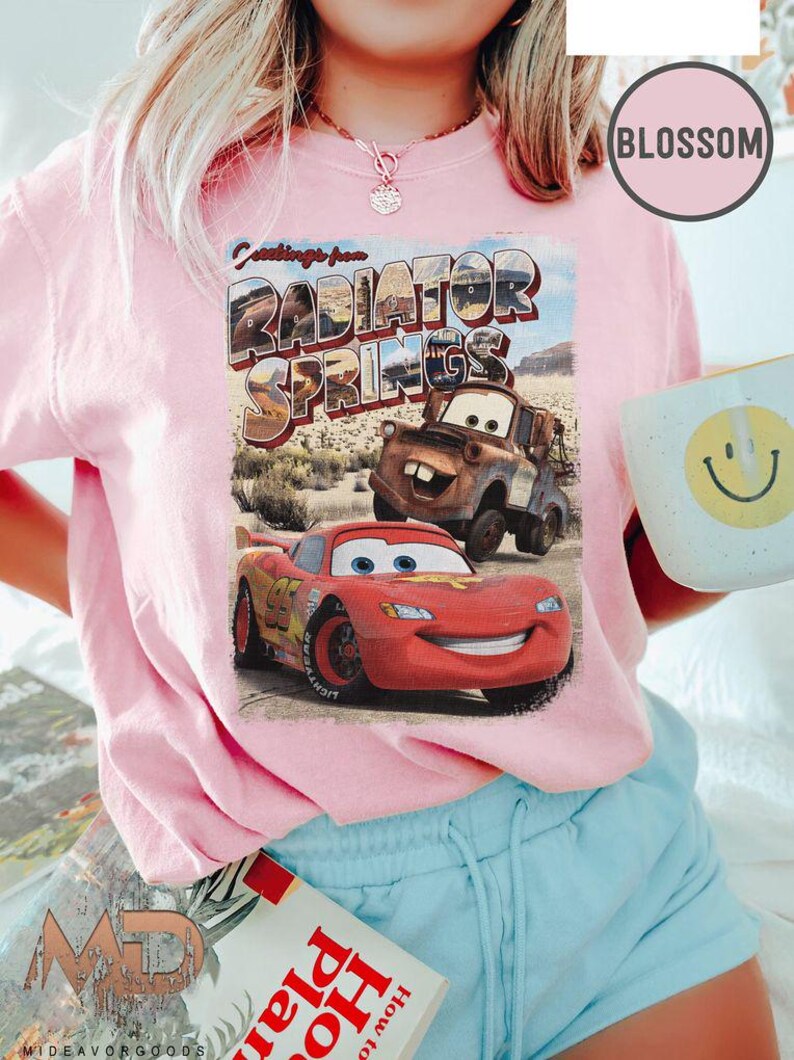 K&ouml;nnte beinhalten: Ein pinkfarbenes T-Shirt mit einem Vintage-Postkarten-Design mit dem Text "Greetings from Radiator Springs". Die Postkarte zeigt ein rotes Rennwagen und einen rostigen Abschleppwagen.