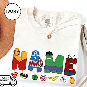May include: Ivory-colored t-shirt with the word "NAME" in colorful superhero-themed letters. The shirt features various superhero logos and a gold necklace. The shirt is made of Comfort Colors material.